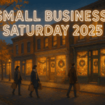 Small-town main street at twilight with glowing Christmas storefronts, golden autumn leaves, and bundled shoppers walking with shopping bags beneath the bright header text “Small Business Saturday 2025.”