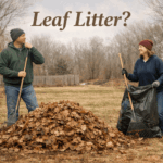 Couple in sweatshirts and knit caps raking a pile of damp brown leaves in an early-spring yard under soft overcast light.