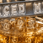 Extreme close-up of a sparkling glass of golden ginger ale filled with crystal-clear ice cubes containing laser-engraved miniature runners on hamster wheels, beneath an ice cube tray spelling “Iced Me” with one empty slot between the words.